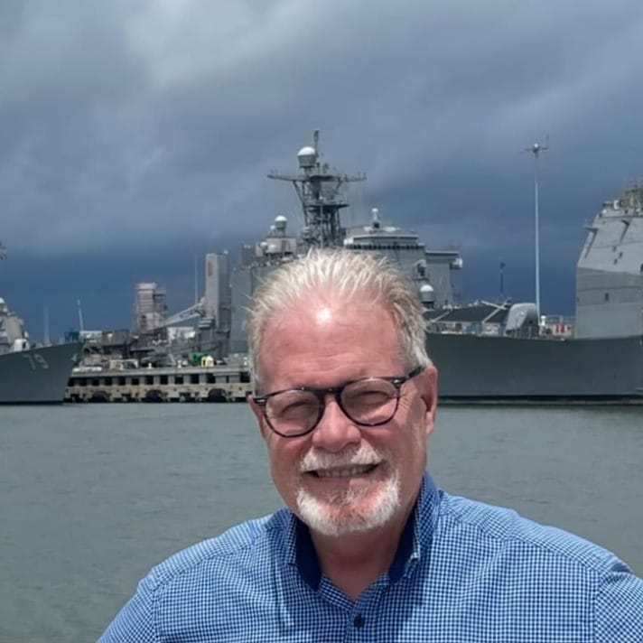 A photograph of Dr. John Cordle with the USS Oscar Austin and USS San Jacinto moored alongside a pier in the background. The sky is cloudy, as if a storm is coming.